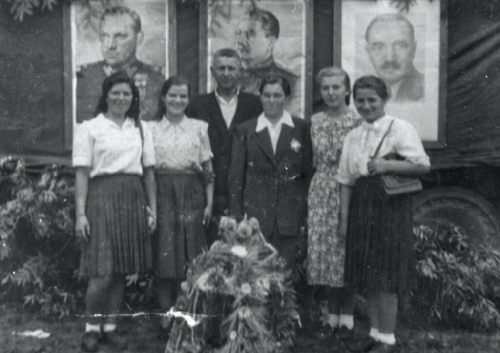 Na czarno-białej fotografii znajduje się grupa sześciu osób pozujących na tle trzech dużych portretów. Portrety przedstawiają mężczyzn w formalnych ubraniach lub mundurach, postaci historyczne i polityczne. Grupa składa się z pięciu kobiet i jednego mężczyzny, ubranych w stroje z połowy XX wieku. Na pierwszym planie widoczna jest wiązanka kwiatów i zbóż ustawiona na ziemi. Tło zdjęcia ozdobione jest roślinnością, a po prawej stronie widoczna jest część pojazdu - ciężarówki.