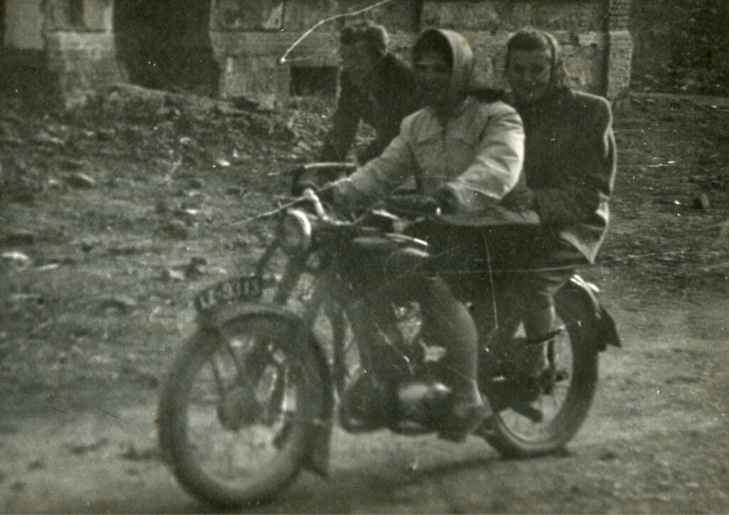 Na czarno-białej fotografii dwie kobiety siedzą na motocyklu i jadą po nierównej drodze w otoczeniu starych budynków. Jedna z kobiet prowadzi motocykl, druga siedzi z tyłu. Uśmiechają się. Za nimi na rowerze jedzie mężczyzna. Zdjęcie ma dynamiczny charakter.